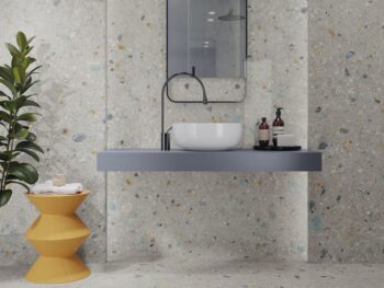 Płytki łazienkowe lastryko - APE Ceppo Fantasy Rect 60x120 cm. Ściana i podłoga w łazience z kolorowymi kaflami lastryko. Biała umywalka, szara półka i pomarańczowy stolik.